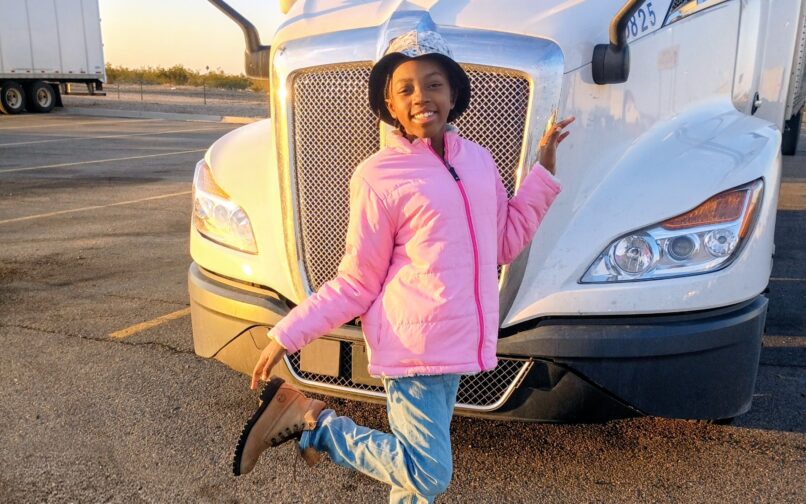 Person wearing a pink jacket, light blue jeans, tan boots, and a hat, posing with one leg raised in front of a white semi-truck in a parking lot during warm, low-angle sunlight.